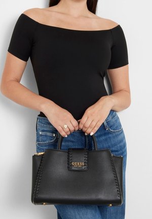 Vrouw in een zwarte off-shoulder top en blauwe jeans die een zwarte Guess-handtas met gevlochten details en gouden logo vasthoudt.
