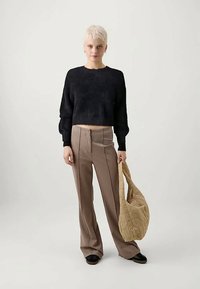 Cilvēks ar īsiem baltiem matiem, kas valkā melnu īsu džemperi, bēšas platleņķa bikses, melnas kurpes un tur lielu bēšas krāsas pārvalku.