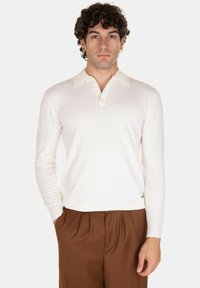 Polo a maniche lunghe color crema realizzato in morbido materiale, con colletto classico e patta con tre bottoni, abbinato a pantaloni marroni.