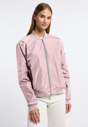 Jonge vrouw die een lichtroze bomberjack draagt, dichtgeritst, gecombineerd met een witte broek, staande tegen een effen witte achtergrond.