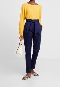 Persona che indossa un maglione giallo, pantaloni a vita alta blu navy con una cintura annodata, sandali con stampa serpente e tiene in mano una piccola borsa bianca con manici in legno.