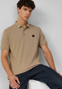 Beige Poloshirt mit klassischem Kragen, drei Knöpfen und kleinem schwarzem Logo auf der Brust; glatte Textur, kurze Ärmel.