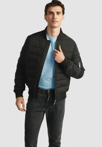 Czarna kurtka puchowa o pikowanej fakturze, z prążkowanymi mankietami i zamkiem błyskawicznym z przodu. Noszona na jasnoniebieskiej koszuli, z detalem bocznej kieszeni.