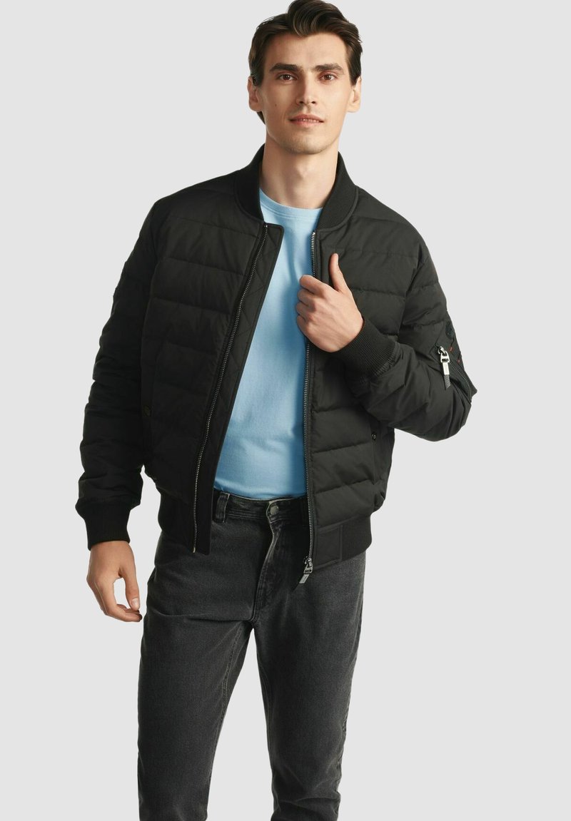 Czarna kurtka puchowa o pikowanej fakturze, z prążkowanymi mankietami i zamkiem błyskawicznym z przodu. Noszona na jasnoniebieskiej koszuli, z detalem bocznej kieszeni.