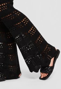 Jambes vêtues d’un pantalon large noir en crochet et de sandales plates en cuir tressé noir sur un sol gris clair.