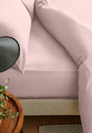 Weiches rosa Bett mit passendem Kissen und Decke, neben einem runden Holztablett mit grünen Blättern, auf beigen und strukturierten Teppichen.