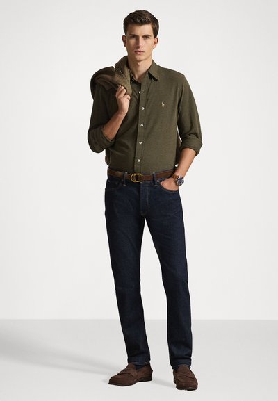 Camicia verde oliva a bottoni, jeans blu navy, cintura marrone e mocassini marrone scuro. Il modello tiene un maglione sulla spalla. Abbigliamento casual.
