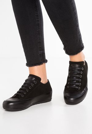 Osoba nosząca czarne, niskie, sznurowane sneakersy z skórzanymi noskami oraz czarne, przycięte dżinsy z postrzępionym wykończeniem na białej podłodze.