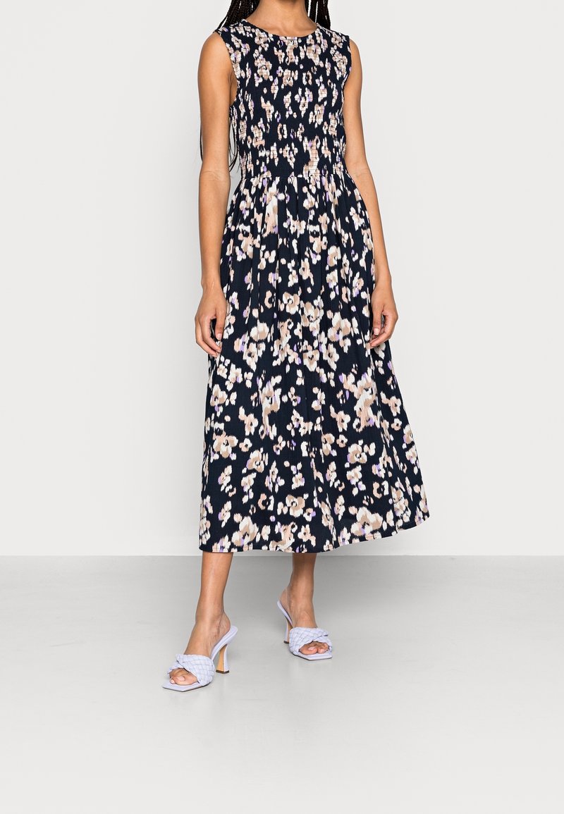Eine Frau steht in einem ärmellosen navyblauen Blumenkleid und hellblauen Absätzen und zeigt eine entspannte Pose vor einem schlichten weißen Hintergrund.