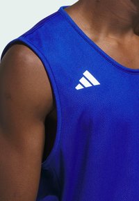 Canottiera sportiva blu in tessuto a rete. Presenta il logo bianco Adidas sulla spalla. Texture liscia con scollo rotondo e senza maniche.