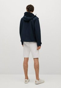 Tmavě modrá bunda s kapucí a lehce volným střihem, se stahovací šňůrkou, nošená přes bílé šortky a světle zbarvené tenisky.