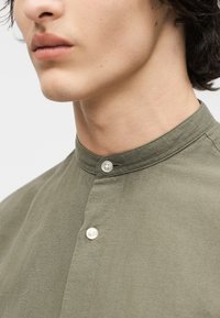 Νεαρό άτομο που φοράει πουκάμισο μανδαρίνι ελιάς με λευκά κουμπιά, εμφανιζόμενο από το πηγούνι μέχρι το πάνω μέρος του στήθους.