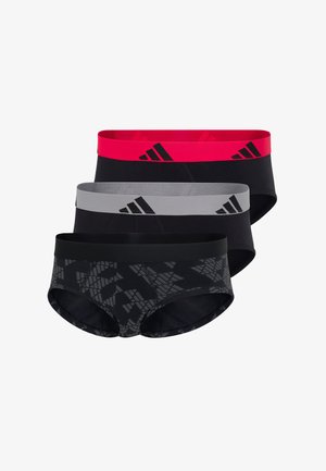 Athletischer Mann steht barfuß da, trägt schwarze Slips mit einem roten elastischen Bund, der schwarze Adidas-Logos zeigt.