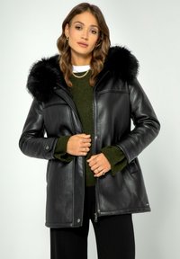 Zwarte faux leren jas met een grote met bont afgewerkte capuchon. Voorzien van een rits en zijzakken. Dragen over een groene trui.