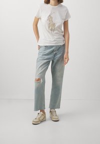 Bílé bavlněné tričko se zlatým motivem hráče póla, doplněné světle modrými džíny s opotřebovaným vzhledem a krémovými teniskami s béžovými doplňky.