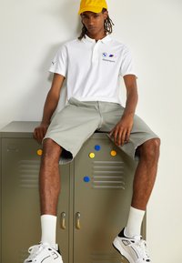 Hvid polo shirt med BMW Motorsports logo, lysegrå shorts og hvide sneakers. Modellen sidder på et metalgrønt skab med farvede prikker.