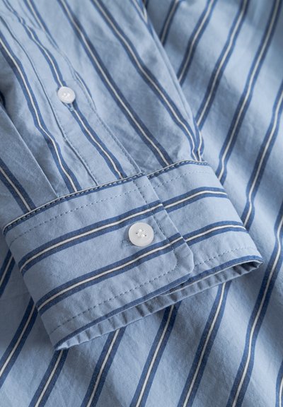 Puño de camisa azul claro con rayas verticales en azul marino, blanco y gris, asegurado con un botón blanco sobre tela a rayas.