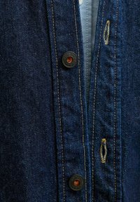 Camicia di denim in tessuto blu scuro, con cuciture arancioni, due bottoni neri e un'asola; presenta uno strato interno azzurro chiaro.