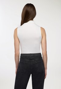 Donna con lunghi capelli castani, che indossa una canotta bianca senza maniche e jeans neri a vita alta con cintura borchiata, in piedi di fronte a uno sfondo bianco uniforme.