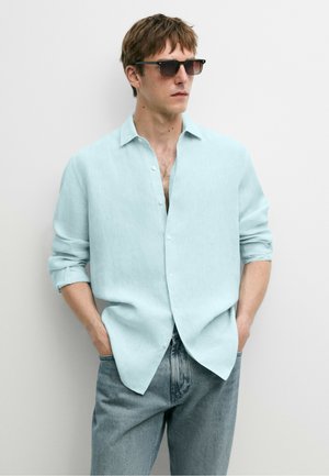 Mann trägt hellblaues Hemd mit hochgekrempelten Ärmeln und Sonnenbrille, steht mit Händen in den Taschen vor einer einfarbigen Wand.