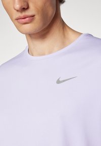 Šviesiai violetinė Nike trumpomis rankovėmis marškinėliai su apvaliu kaklu; priekyje yra pilka Nike logo. Audinys atrodo tekstūruotas.