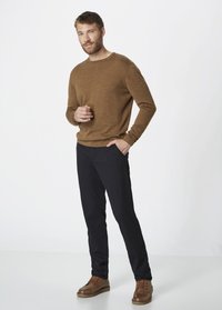 Brauner Wollpullover, marineblaue Hose und braune Schnürschuhe. Das Modell steht mit der Hand in der Tasche und zeigt eine entspannte Passform sowie neutrale Farben.