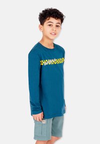 Langärmliges blaues Baumwoll-T-Shirt mit einem gelb-schwarzen Schachbrettmuster und dem Text "find & Rascal." Kombiniert mit grünen Shorts.