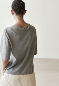 Osoba o ciemnych włosach związanych w kok, ubrana w luźną szarą bluzkę z krótkim rękawem i jasną spódnicę, stojąca tyłem do kamery.