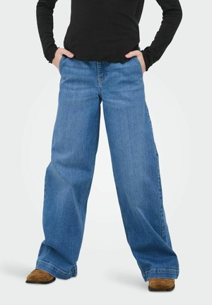 Persona con pantalones vaqueros anchos azules y las manos en los bolsillos, combinados con una camiseta de manga larga negra y zapatos marrones.