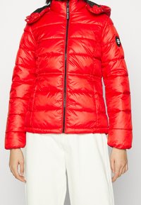 Röd puffjacka med dragkedja framtill, svarta detaljer på kragen och ett quiltat mönster. Har en tajt passform och en uppstående krage.