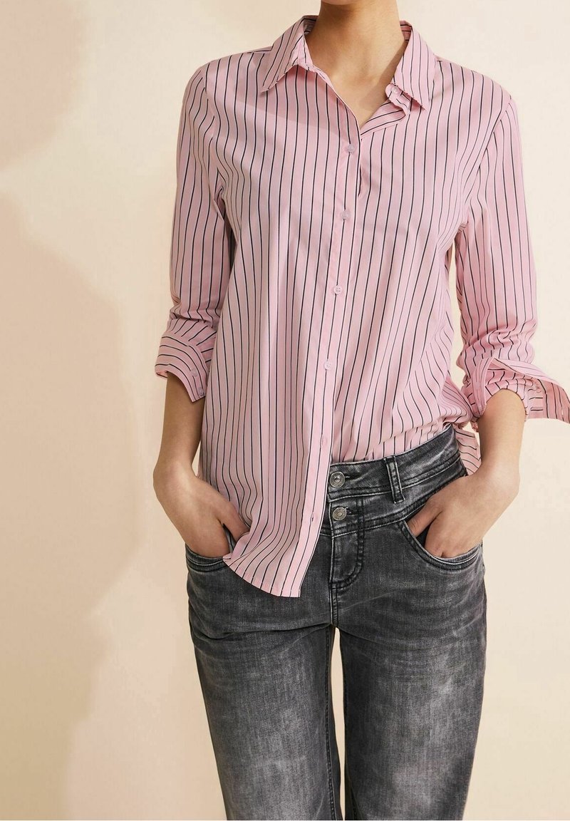 Personne portant une chemise rose à manches longues avec des rayures verticales noires et un jean noir délavé, les mains dans les poches.