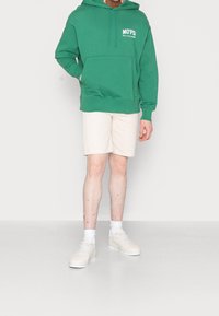 Sudadera verde con bolsillo frontal y logo blanco, combinada con pantalones cortos beige y zapatillas blancas. Tejido suave, corte relajado.