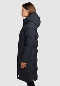 Zwarte pufferjas met capuchon, voorzien van een ritszak aan de zijkant, een geborduurd ontwerp en een middellange snit. Soepele, matte textuur.