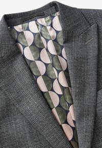 Blazer grigio con texture e fodera a motivi, caratterizzata da grandi cerchi verde e rosa, e strisce alternate, che mostrano colori contrastanti.