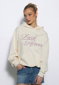 Kremowa bluza z kapturem, z przednią kieszenią, z haftowanym napisem "Local Heroes" w różowym stylu. W zestawieniu z dżinsową spódnicą.