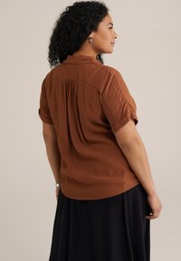 Blusa a maniche corte di colore ruggine con motivo testurizzato, caratterizzata da un retro plissettato e un colletto. Abbinata a una gonna nera.