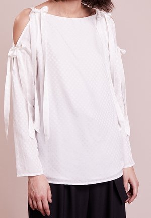 Hvid bluse med lange ærmer, kolde skulderudskæringer og dekorative skulderbånd, båret med sorte bukser på en lys baggrund.