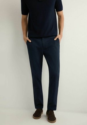 Mężczyzna w granatowej koszuli z krótkim rękawem i dopasowanych spodniach w tym samym kolorze, noszący brązowe zamszowe mokasyny, stoi z rękami w kieszeniach.