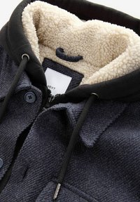 Námornícky modrá bunda s čiernou kapucňou, so zipsovým zapínaním a veľkými gombíkmi. Podšitá krémovou fleecovou podšívkou pre dodatočné teplo a pohodlie.