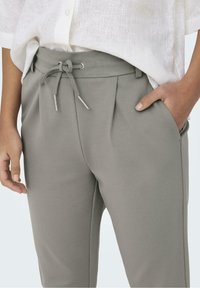 Pantalons gris en tissu lisse avec taille à cordon, dotés de plis sur le devant et de poches latérales. Associés à une chemise en lin blanche.