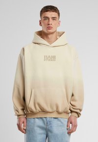 Beige, oversized hoodie med känguruficka, ribbade �ärmslut och nederkant. Har en subtil "KANI STUDIOS"-logotyp i en ljusare nyans.