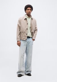 Άνδρας με αφροχτένισμα, φοράει μπεζ σακάκι, ανοιχτό πράσινο πουκάμισο με ρίγες, φαρδιά ανοιχτό μπλε τζιν και μαύρα παπούτσια, στέκεται με το ένα χέρι στην τσέπη.