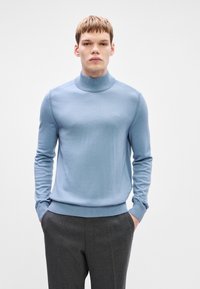 Ανοιχτό μπλε πλεκτό πουλόβερ με ψηλό λαιμό, εφαρμοστή σχεδίαση, ριμπ μανίκια και ζώνη μέσης. Διαθέτει μικρό κεντημένο λογότυπο στο στήθος.