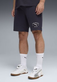 Shorts de sport bleu marine à taille élastique, ornés du logo blanc "PUMA ATHLETICS". Portés avec des chaussures blanches et des chaussettes blanches avec des accents de logo noir.
