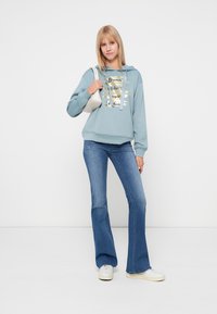 Hanorac bleu deschis cu text grafic auriu, asortat cu blugi evazați albaștri și adidași albi. Design simplu și casual, cu o croială relaxată.
