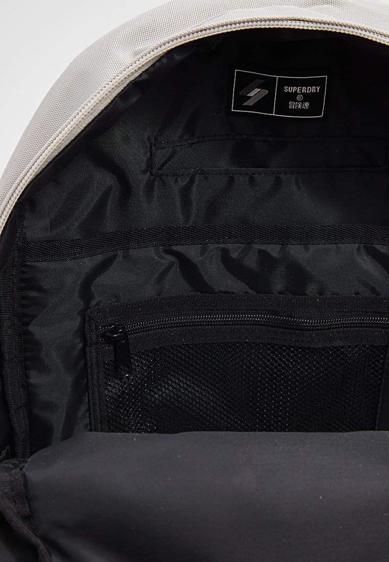 Open beige en zwarte rugzak met zichtbare binnenkant, inclusief een mesh ritsvak en een Superdry merklabel.