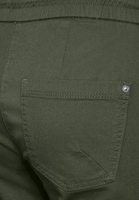 Pantalons olive en tissu uni, présentant une poche arrière avec un rabat pointu et un accent en métal argenté. Détails de couture visibles.
