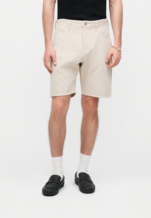 Mężczyzna noszący beżowe szorty do kolan, białe skarpetki, czarne mokasyny i czarną koszulę, stojący na jednolitym jasnym tle.