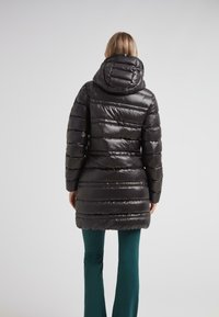 Mujer vestida con un abrigo acolchado negro brillante con capucha, de espaldas, combinado con pantalones ajustados verde oscuro.