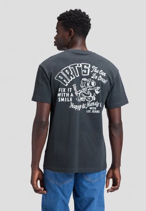 Νέος άνδρας που φοράει μαύρο T-shirt με λευκό γραφικό και κείμενο που δείχνει καρτούν τεχνίτη επισκευής και φράσεις για να διορθώνεις με χαμόγελο.